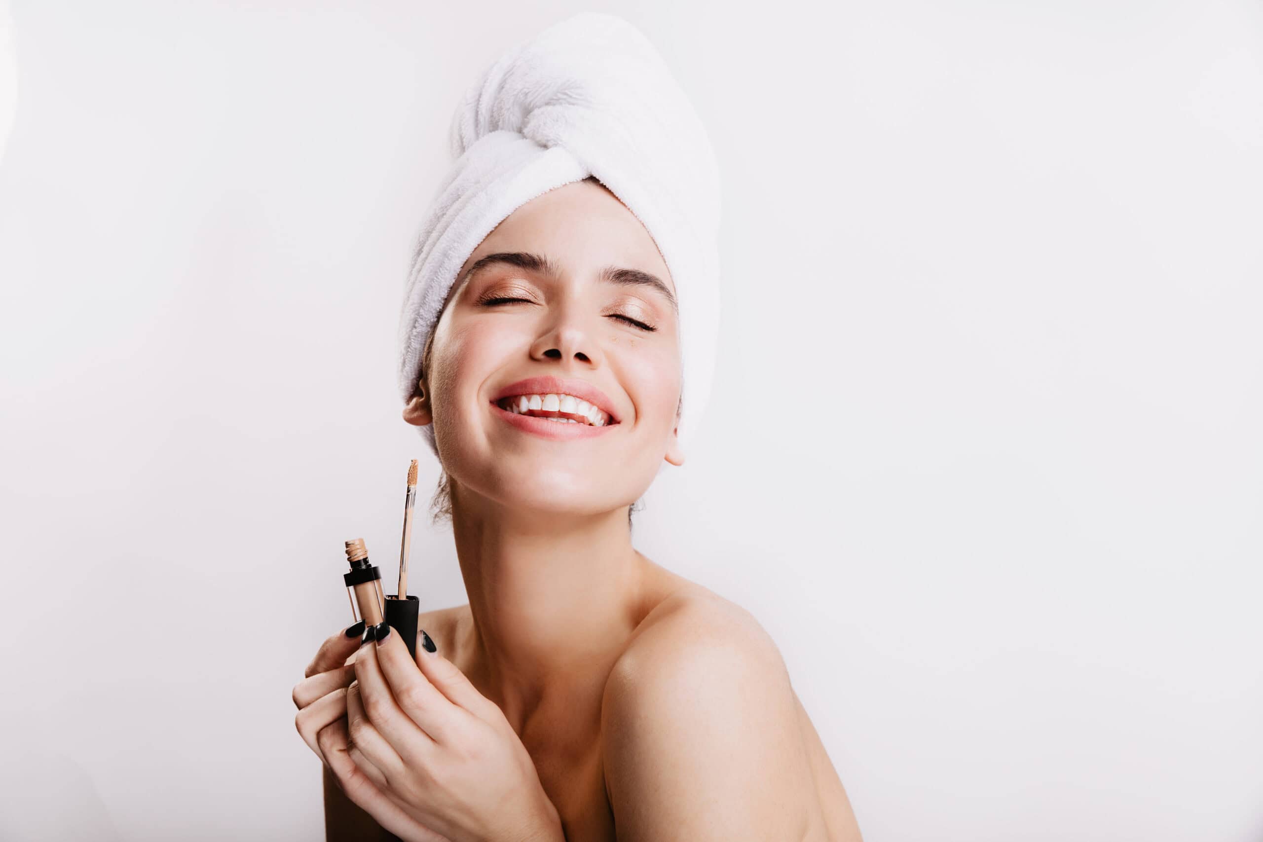 Girl with snow white smile and closed eyes poses on white background. Model with perfect face tone holds concealer.