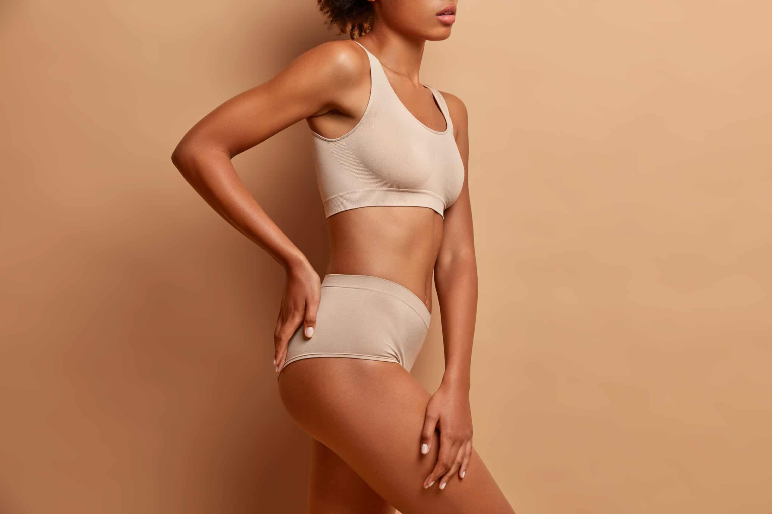 Sexy body of mixed race woman standing in profile keeps hand on buttocks dressed in underwear has perfect shining skin slim figure